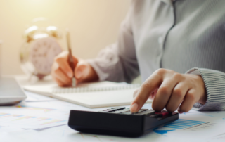 Person using a calculator and writing in a notebook at a desk with financial documents, suggesting tax planning or financial review.